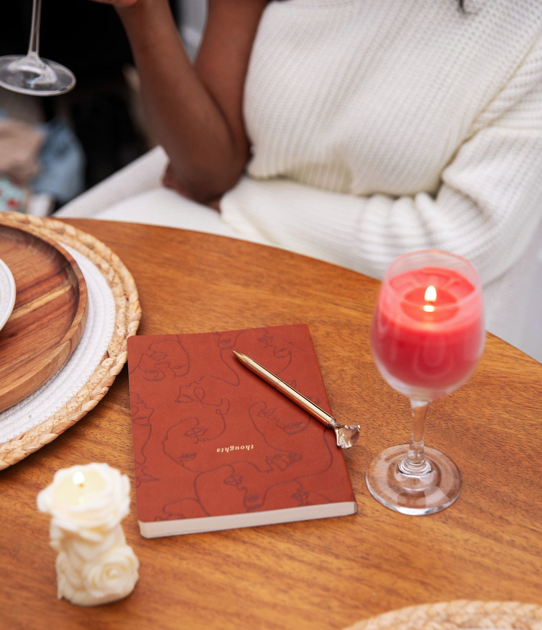 Red notebook with a pen on a wooden table next to a lit candle in a glass holder.